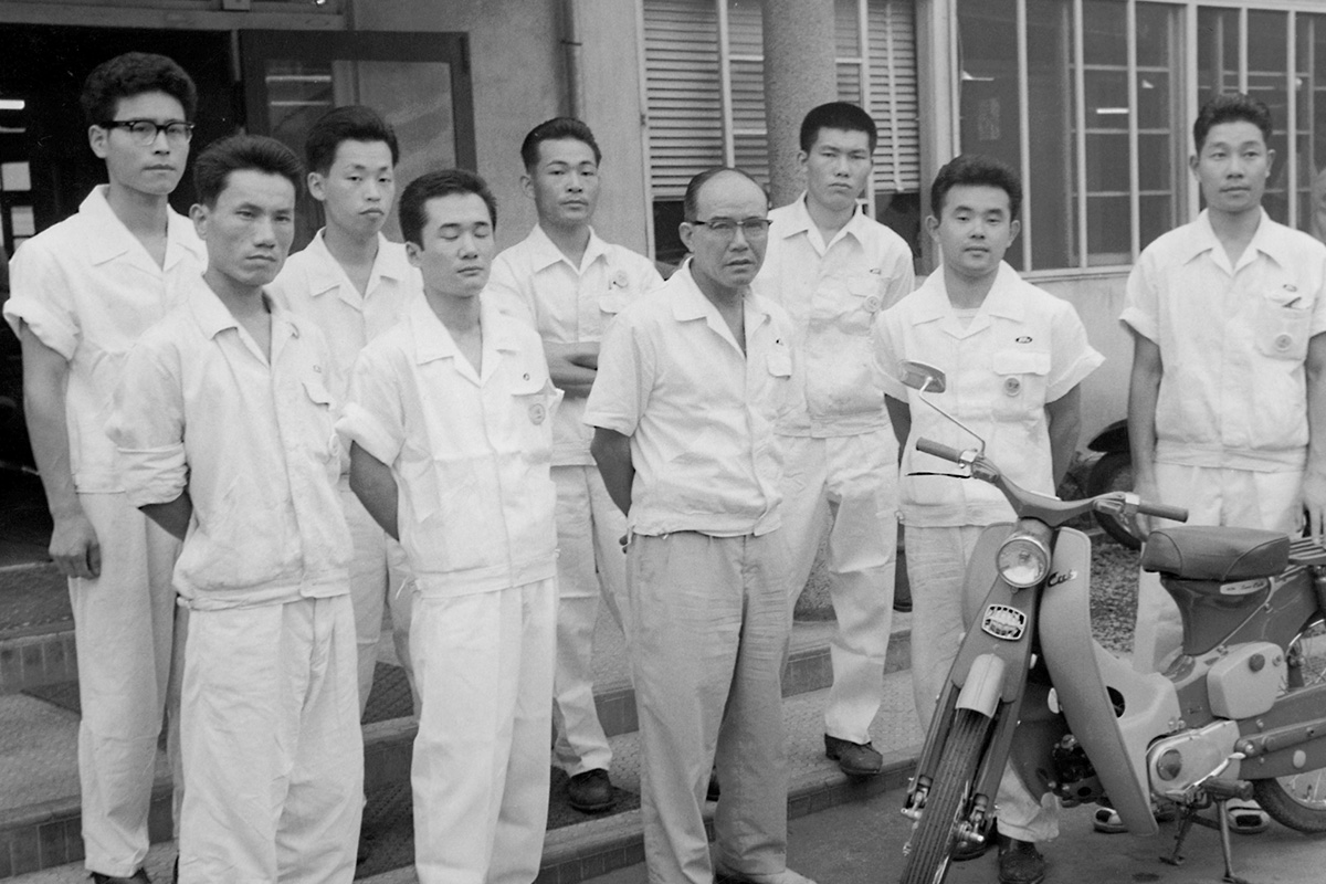 Soichiro Honda with the Super Cub's development team. After many months of trial and error and ”waigaya” (’boisterous meetings'), a unique machine unlike anything seen before was born.