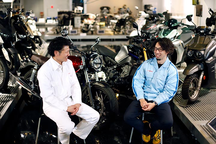 Test riders Sasazawa (left) and Yamada (right) smile as they are surrounded by motorcycles.